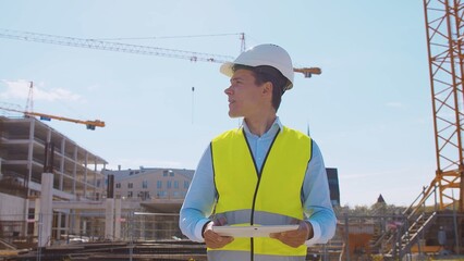 Professional builder standing in front of construction site. Foreman in hardhat helmet and vest. Office building and crane background. Business, real estate and investment.