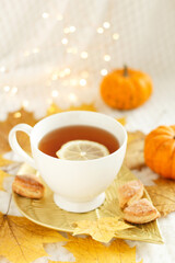 Creative mood. A cup of tea with lemon, cookies, autumn leaves and pumpkin on a white plaid with lights. Cozy autumn composition. Autumn mood concept.