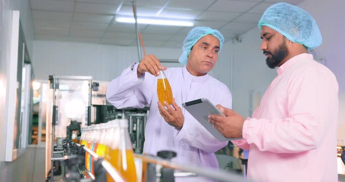 In An Industrial Water Factory, A Food Scientist Worker Wearing A Hairnet And A Face Mask Converses While Monitoring Quality Control And Putting Comments On Juice Bottles. Beverage Line Inspection