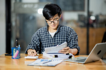 A young financial market analyst works in the office with his laptop while sitting at a wooden table. businessman analyzing graph document in hand.