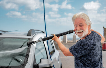 Smiling caucasian senior man washing his car in a self-service car wash station using high pressure...
