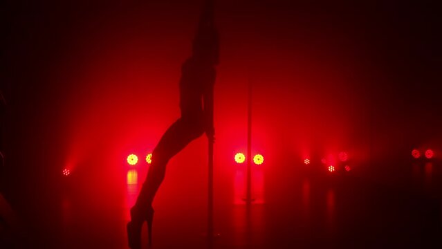 Slim woman performing poledance tricks sensually in spotlights. Sexy lady making moves upside down in night club. Young blonde dancer doing twine on pylon illuminated soft nightclub backlight.