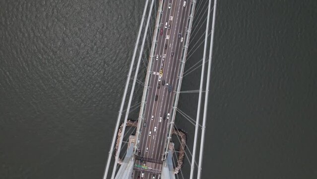 A high angle view over New York Harbor and the Verrazano Bridge during the day. The camera tilted down, dolly out and pan left high over one of the tall towers to look down on traffic on the span.