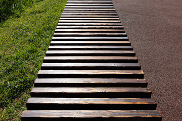 A modern long bench made of worn wooden slats looking into perspective. Separates the sidewalk and the green lawn. Selective focus.