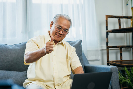 Portrait of smiling asian senior mature man sitting on couch sofa using laptop show thumbs up for easy technology, happy senior old male recommend good quality working on computer at home.