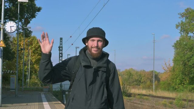 Young man with a beard waving his hands in greeting at the railway station
