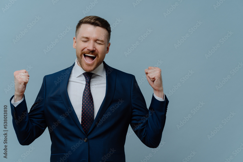 Canvas Prints Overjoyed triumphing businessman clenches fists with triumph celebrates successful deal dressed in formal clothes isolated over blue background copy space for your advertisement exclaims from joy