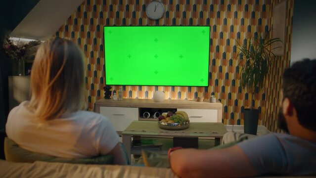 Couple sitting on sofa in living room, talking, watching breaking news, movie or series on TV. Wife and husband resting and chilling at home in the evening together. Green screen. Chromakey.
