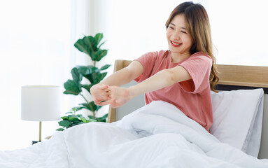 Millennial Asian young happy cheerful lazy sleepy female teenager sitting laying smiling on pillow under thick warm blanket on cozy bed in bedroom at home stretching arms after waking up in morning