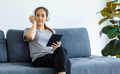 Millennial Asian female housewife sitting on cozy sofa wearing wireless stereo headphone set holding touchscreen tablet computer listening to streaming online music playlist in living room at home