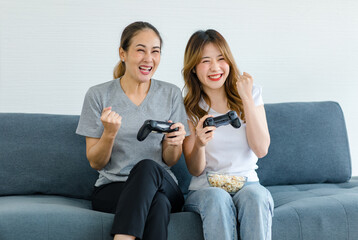 Millennial Asian young happy cheerful joyful female lesbian LGBTQ lover couple sitting smiling holding joystick controller playing winning victory online streaming game together at home living room