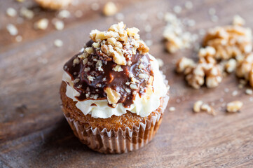 Tasty chocolate muffin with creme mascarpone and chocolate on top, covered with nuts. Placed on wooden desk. Sweet fresh and homemade cupcake.