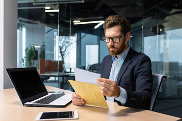 Serious and focused mature boss working inside modern office building with laptop, businessman in business suit carefully reading letter from bank, upset and disappointed man in glasses and beard.