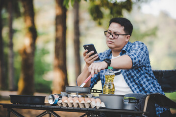 Portrait of Asian traveler man glasses using smartphone while cooking a meal at a campsite