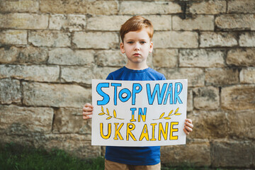 Sad boy holding poster in colors of national flag with words Stop War In Ukraine against brick wall...