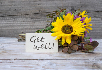 Card with the English text: Get well lies in front of an autumn bouquet of flowers with a sunflower.