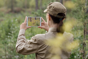 Woman biologist in uniform take a photo using a digital tablet in a national park in summer, back...