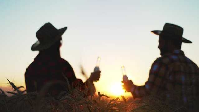Silhouette two people farmers in field drinking wheat beer of natural production after harvesting. Concept of brewing at field nature landscape background with sunset in sunbeams. Product tasting.