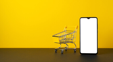 Cyber monday concept. Photo of smartphone and empty shopping cart on tabletop yellow wall...