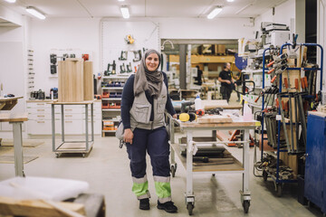 Full length portrait of smiling female carpenter standing by workbench in workshop