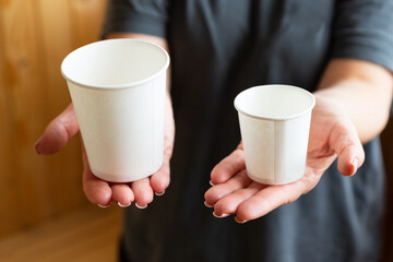 Large and small paper cups on the palms of female hands. Large and small for comparison. Offers of various sizes. Ecological paper tableware.