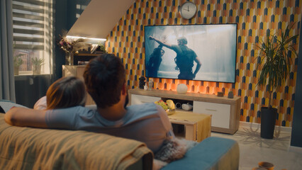 Couple sitting and hugging on sofa in cozy living room, watching action movie on TV or criminal...