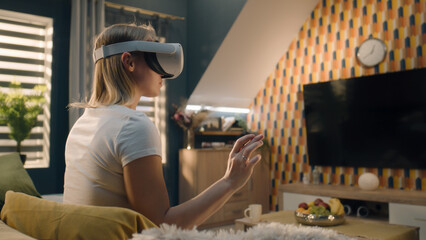 Woman wearing VR headset, sitting on sofa in cozy living room in midday, gesturing, playing video game in virtual reality, testing VR goggles, spending weekend at home. Cyberspace and metaverse.