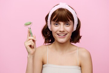 An emotional woman with clean skin, an ideal smile and dark beautiful hair stands on a pink background with a beautiful bandage on her head with a facial massage roller.Horizontal studio shot.