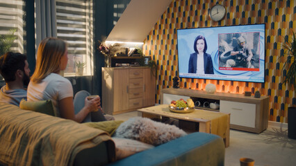 Couple sitting on sofa in living room together, talking, watching TV news about war in Ukraine, Armenia, Syria. Man and woman spending time on weekend at home. TV live broadcasting.