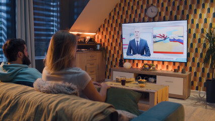 Couple sitting on sofa in living room with snacks, watching TV news about rivalry between Russian and United states, cold war. Man and woman spending evening at home. TV live broadcasting.