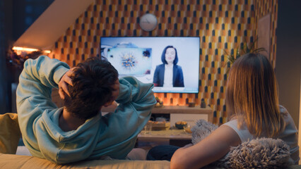 Couple sitting on sofa in living room, looking at each other, watching TV news about poliovirus and...
