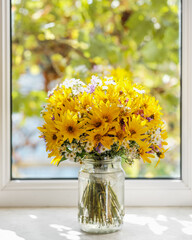 Bouquet of yellow, violet and white wild flowers in a jar against window. Autumn composition.