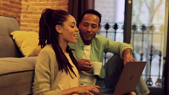 Happy young black man and woman sitting on the floor in the apartment, watching movie online or looking for good trip, planning vacation, copy space