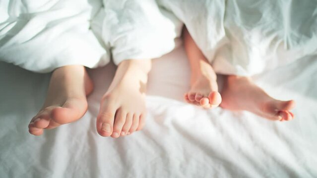 Close-up of bare feet of mother and child from under white blanket on bedding. Parent and newborn lie in bed. Love and care in happy family. New life and parenthood. Sun rays at living room indoors.