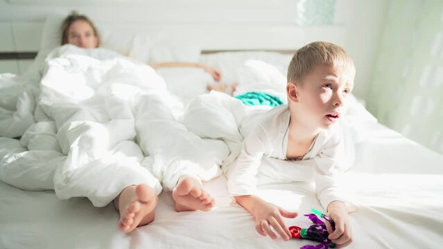 Home life brother and sister or mother in bedroom lie on bed with white linens. Sun rays in home interior in morning. Baby boy is playing with transformer robot toy. Family happy leisure activity.