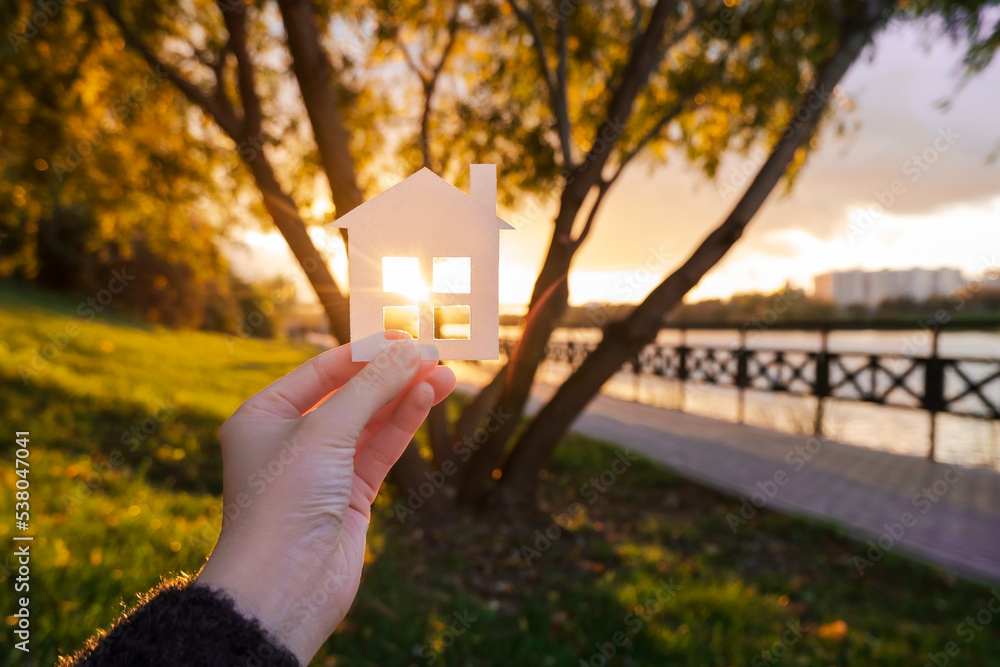 Wall mural Hand holds a paper house against the background of the sunset sky. The sun is setting over the horizon. The concept of your own home, personal space and mortgage