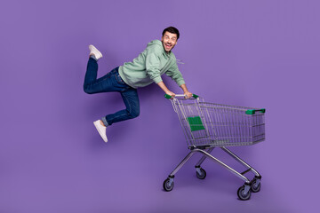 Full body portrait of crazy carefree person jump fly push shopping trolley isolated on purple color...