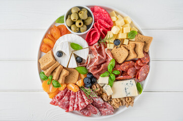 Charcuterie board with a variety of cheeses, salami, chorizzo, prosciutto, honey, grapes, nuts, olives, bread, blueberries and fresh herbs on a white wooden background. A festive snack.