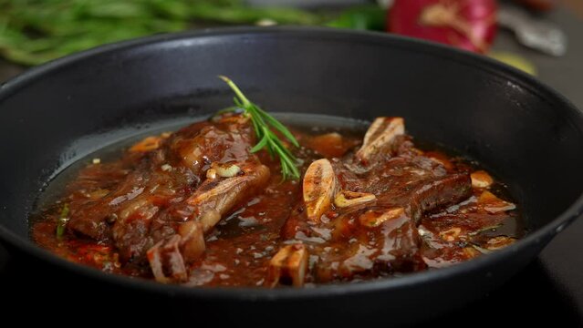 Close up of steak beef ribs in sizzling sauce with garlic fry in hot pan on stove. Hand puts greens rosemary in cooking pieces meat on bone. Food concept