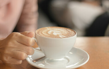 Closeup image of a hand holding a cup of hot latte coffee with blurry room on table at cafe coffee shop.