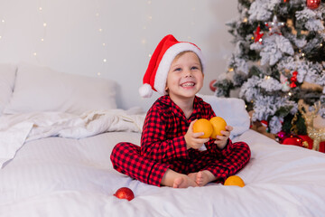 happy little boy in pajamas and Santa hat at home in bed eating tangerines for Christmas