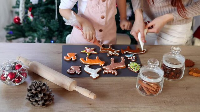 Happy little girl with her beautiful mom coloring Christmas gingerbread cookies at home. Christmas decoration in the kitchen. Fir tree with fairy lights. The concept of the New Year and Christmas. Hol