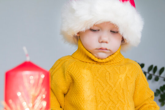 A cute little boy in a yellow sweater and a red Santa Claus hat, with a red Christmas candle. Celebrate Christmas.