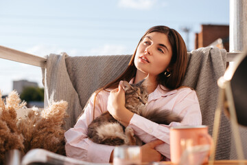 A woman with long hair in pink pajamas with a Maine Coon cat in her hands drinks coffee in the...