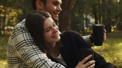 Young lady cuddles up to the chest of her boyfriend and drinks takeaway tea. Sweet family couple sitting on the grass outdoors in a warm embrace, talking about their plans, hopes and dreams