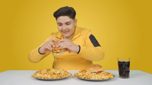 Unhealthy foods, obesity problem. Orange background.
Obese boy eating hamburger with pleasure.

