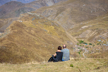 Fototapeta premium A man and a girl are sitting on a hill in the mountains. Happy couple. The man hugs the girl. Caucasus mountains, Ossetia.