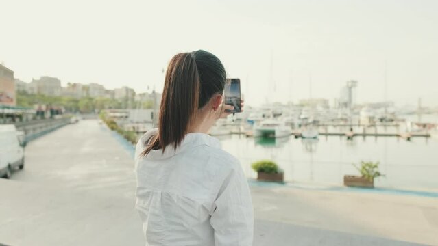 Young woman taking photo of the sea port on mobile phone. Back view