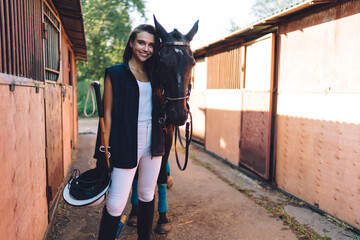 Positive horsewoman embracing stallion near stables in countryside