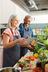 Happy senior diverse couple wearing aprons and using tablet in kitchen